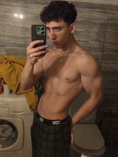 A young male individual is depicted in a mirror selfie taken indoors. He possesses a muscular build with visible abdominal muscles and defined biceps. His dark, curly hair is styled, and he looks directly at the camera with a neutral expression. He is shirtless, wearing only dark green plaid pajama-style pants featuring a black waistband with "LEVI'S" branding. His left hand, viewed from the subject's perspective, holds a dark-colored smartphone with a visible multi-lens camera array, used to capture the mirror reflection. His right hand rests on his hip. The background elements include a white front-loading washing machine to the left, partially covered by a crumpled yellow garment. To the right, a white toilet with a closed lid is visible. The room's walls are tiled with light-colored, horizontally patterned material. The specific location within As-Salihiya Municipality, Syria, cannot be determined from the image's visual context.
