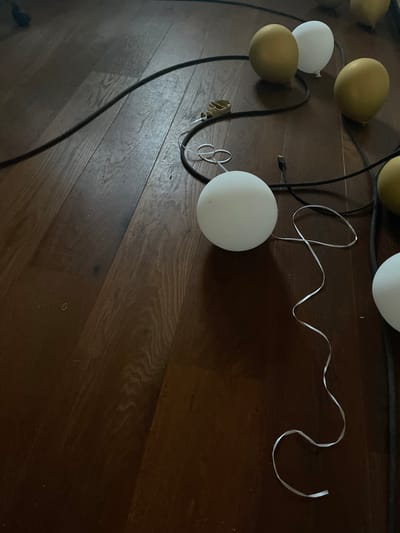 The image depicts a close-up, overhead view of several gold and white balloons on a dark wooden floor. A black cable is visible, weaving among the balloons, and a white ribbon curls on the floor next to one of the white balloons. There are no people or identifiable landmarks of Ореховка, Russia present in the image. The scene appears to be indoors, with the focus solely on the balloons and the floor.