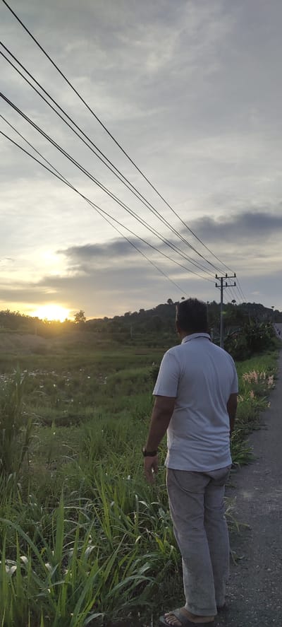 The image shows a man standing on a road in Seuneubok, Indonesia, looking out at a field at sunset. The field is covered with tall green grass and some scattered vegetation. In the background, a low hill is visible. Power lines stretch across the sky. The man is wearing a light-colored polo shirt, grey pants, and sandals. He appears to be wearing a watch on his left wrist. The road is paved and runs along the edge of the field. The sky is cloudy with the sun setting in the distance.