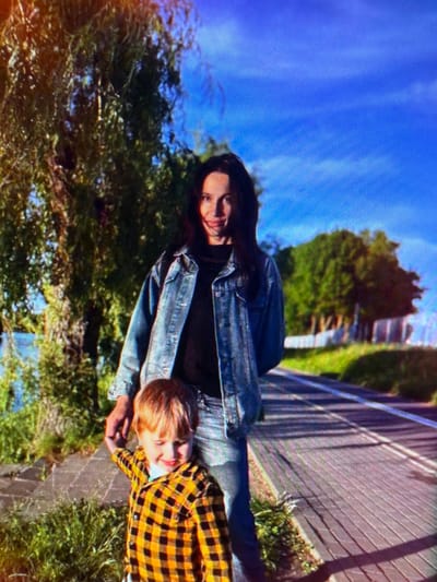 A woman and a young child are standing on a paved pathway next to a body of water, possibly a river, with trees and lush green vegetation on either side. The woman, with long dark hair, is wearing a blue denim jacket and jeans, and appears to be smiling. The child, in a yellow and black checkered shirt, is holding the woman's hand and looking down with a slight smile. The pathway has a distinct pattern of wooden planks and is bordered by grass. The sky is a clear, bright blue, suggesting it is a sunny day. The location is identified as Папярнянскі сельскі Савет, Belarus, which is a rural administrative division.
