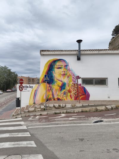 The image shows a street scene in Dénia, Spain, featuring a building with a large, colorful mural of a woman's face. The mural, painted on a white-walled building, displays a woman with vibrant colors and multiple layers, with visible tattoos and jewelry. A pedestrian crossing sign is visible in front of the mural.
The building has a tiled roof with a black chimney. A window with a metal grate is to the right of the mural. To the left of the building is a street with a "STOP" sign and a "No Right Turn" sign, along with a trash can. Cars are parked along the side of the road. The sky is overcast with grey clouds. A crosswalk is in the foreground. A bike lane is between the sidewalk and the street.