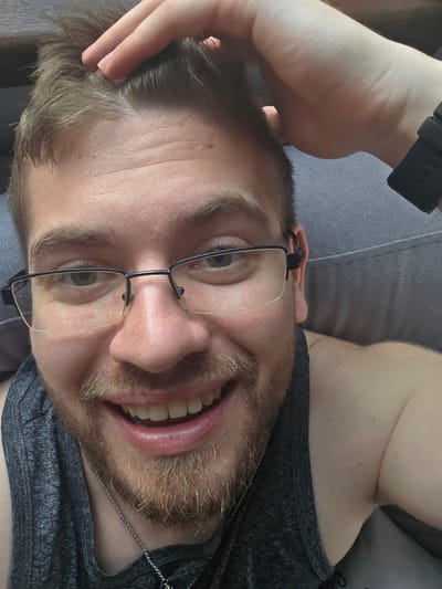 The image presents a close-up, angled portrait of a young adult male lying down indoors, likely in Yagodovo, Bulgaria. He is positioned with his head tilted towards the left of the frame, looking directly at the camera with a wide, open-mouthed smile, revealing white teeth. The man has light brown hair, a light beard or stubble, and green or hazel eyes. He is wearing thin, black-framed glasses.
He is dressed in a dark grey, heathered sleeveless top or t-shirt, and a thin, metallic ball-chain necklace is visible around his neck. His left hand is raised, with fingers lightly touching his hair on the side of his head. A black digital wristwatch is worn on his left wrist. The setting appears to be a comfortable indoor space, indicated by the grey upholstered surface, possibly a couch or bed, on which he is resting. The lighting is bright and diffused, suggesting it is daytime. The man's expression conveys happiness and relaxation. Small red spots are visible on his forehead. No visible text is present in the image.