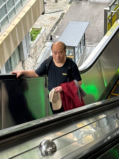 A man is riding an escalator. He is holding a hat and a jacket. The escalator is green. The location appears to be the Nan'an District of China, based on the proximity of the escalator to buildings and street-level scenery.