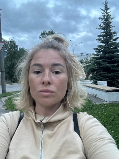 A medium shot shows a woman with blonde hair pulled back in a messy bun and wearing a light beige hooded sweatshirt with a zippered front. She is standing outdoors with a cloudy sky above. Behind her, to the left, is a traffic sign and a utility pole with wires. Green grass and some trees are visible. To the right, a tall evergreen tree stands next to a white structure with a wooden bench. Buildings and a street are visible in the background, suggesting an urban or suburban setting. The image appears to have been taken in Vologda, Russia.
