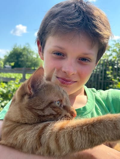A young boy with light brown hair and freckles is holding an orange tabby cat. The boy is smiling and looking at the camera, while the cat is looking to the side. They are both in an outdoor setting, with a blue sky and some greenery in the background. The image appears to be a selfie, taken on a sunny day. There is no indication of the location in the city of Уральское, Russia.