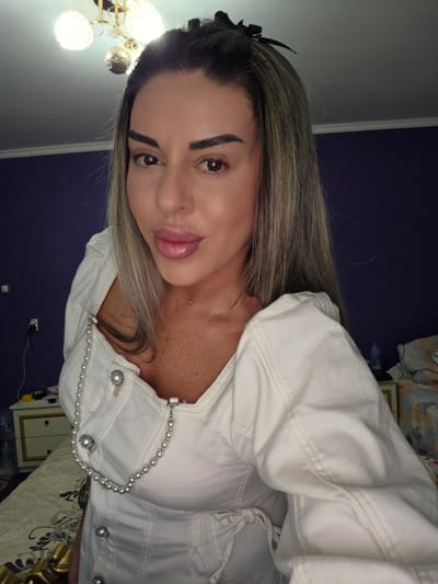 A blonde woman with heavy makeup, wearing a white corset-like top with pearl embellishments, poses for a selfie indoors. The background includes a purple wall, a dresser with drawers, a light fixture on the ceiling, and a bed. The image does not provide any information about the city of Peshtera, Bulgaria.