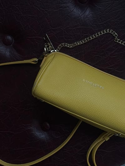 The image shows a yellow Lancaster brand purse with a silver chain lying on a dark brown leather surface, likely part of furniture such as a chair or ottoman based on the tufted design. The word "LANCASTER PARIS" is visible on the purse. The location is unknown and cannot be determined as Zovuni, Armenia based on the image. There are no people visible.