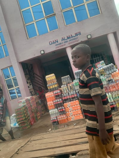 Here is a detailed description of the image:
**Setting:** The image appears to be taken at a small retail shop in Kafanchan B, Nigeria. The shop, identified by the signage "DAN ALMAJIRI," sells provisions and general goods.
**People:**
* A young boy wearing a striped shirt stands near the shop's merchandise.
* Partial views of other individuals are present near the bottom of the image.
**Objects and Merchandise:**
* Stacks of bottled drinks, including Maltina, are prominently displayed in front of the shop.
* Other food items, possibly noodles or similar products, are stacked towards the lower part of the frame.
* The shopfront features windows with a grid pattern.
* The exterior is painted in light pink.
**Actions and Interactions:** The boy's presence indicates a potential transaction or his involvement with the shop. The overall scene suggests a typical small business environment in a residential area.
**Environment:** The ground in front of the shop is wooden planks.