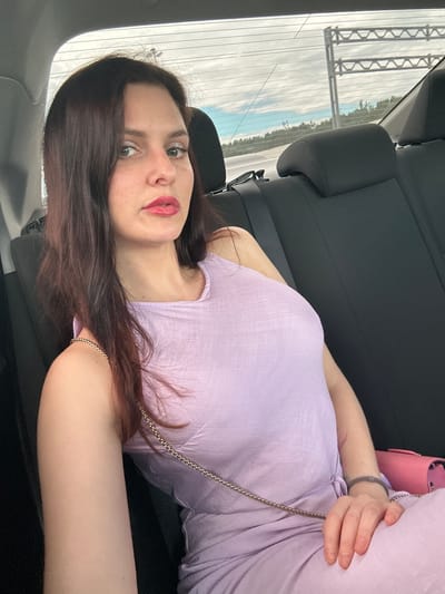 A woman is taking a selfie in the back seat of a car. She has dark brown hair and is wearing a light purple dress. Her lips are painted red. She is holding a small pink purse. The car appears to be on a highway, as indicated by the road signs visible through the rear window. The sky is cloudy. The image is a close-up of the woman, with the background out of focus.