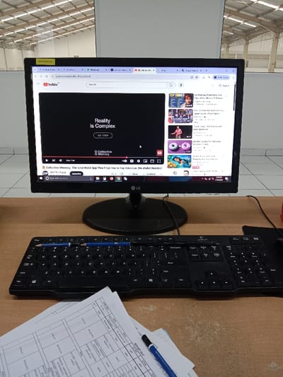 The image shows a desk with a computer monitor, a keyboard, and some paperwork. The computer monitor is displaying a YouTube video with the title "Reality is Complex". The keyboard is black and appears to be a standard computer keyboard. The paperwork is white and has various columns and rows. A blue pen is lying on the paperwork. The scene is set in an office or workspace.  The location is Kecamatan Ngaliyan, Indonesia, based on the provided coordinates.  The workspace is clean and organized, and the environment is bright and well-lit.  The person is not present. The person was likely at the desk, using the computer. The workspace appears to be tidy and there are no other objects present. The scene is peaceful and suggests a sense of work and productivity.  The image appears to be taken with a phone camera.
