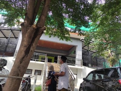 The image, rotated for correct viewing, depicts an outdoor scene in what appears to be an urban or semi-urban environment.
**Subjects:** Two people are visible near a building. One man, positioned to the left of the frame (from the viewer's perspective), wears a white long-sleeved shirt, dark trousers or a sarong, and a dark cap (possibly a peci/songkok). He is looking downwards. A second person, partially obscured behind him, appears to be wearing a dark top and carrying a reddish bag. A black car is parked on the far left, and a white scooter or motorbike is parked on the far right. A prominent tree trunk stands in the right foreground, with green foliage from other trees visible on the left and bottom of the frame, and some in the background. In the midground, there is a multi-story building featuring white and grey walls, balconies with metal railings, some windows, a section with a bright green wall on a lower level, and a visible terracotta-tiled roof on one part.
**Activity or event taking place:** The people are standing near the building, but their specific activity or interaction is unclear. The man appears to be looking down, possibly at something on the ground or in his hand.
**Notable details:** The scene is brightly lit, indicating daytime and sunny weather, with clear shadows cast by objects and people. The architecture of the building, including the balconies and roof tiles, along with the headwear of the man (resembling a peci/songkok), aligns with the provided location context of Kecamatan Asembagus, Indonesia. The image is taken from a slightly low angle.
**Visible text:** No discernible text is clearly visible on any objects or surfaces.