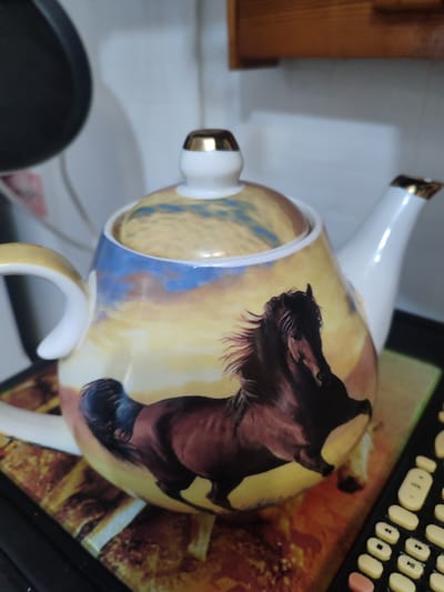 A close-up, slightly angled shot features a ceramic teapot with a dramatic depiction of a dark horse running across a sunset sky. The teapot has a white lid with a gold accent, a white handle, and a white spout with a gold rim. The artwork on the teapot is vibrant, with shades of orange, yellow, and blue representing the sky, and the horse is rendered in dark browns and blacks. The teapot is placed on what appears to be a placemat with a similar sunset and horse motif. In the foreground, to the right, a portion of a black remote control with light-colored buttons is visible. The background is out of focus, showing a white wall and a glimpse of dark kitchen cabinetry. The image was taken in Tuapse, Russia.
