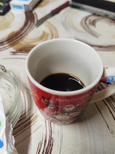 A close-up, eye-level shot shows a red Christmas-themed mug filled with dark coffee. The mug is positioned on a table with a light-colored, patterned tablecloth. To the left of the mug, a portion of a clear glass ashtray is visible, containing a white paper napkin with blue and green markings. In the background, out of focus, are electronic devices and a knife with a red handle. The scene appears to be indoors, likely in a domestic setting in Lomets, Bulgaria, though this specific detail cannot be confirmed from the visual information alone.