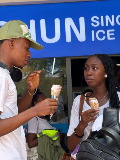A woman and a man are eating ice cream cones in front of a shop in Itirin, Nigeria. The woman has braided hair and is wearing a white shirt, a necklace, and a black bracelet. She is holding an ice cream cone in her right hand and looking at the man. The man is wearing a green cap, a white shirt, and has headphones around his neck. He is holding an ice cream cone in his left hand. There is a sign in the background that says "HUN ICE SINCE". There is a large black bag behind the woman. There is a person in the background wearing a white shirt and a yellow safety vest.