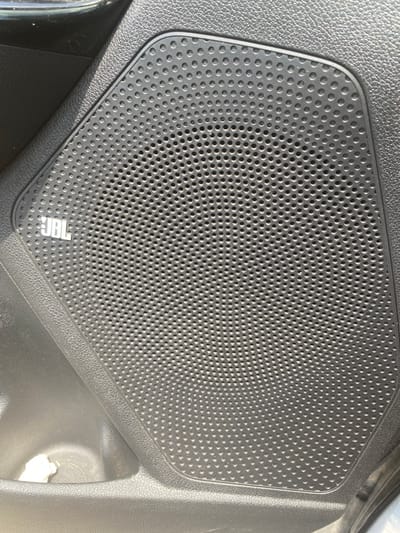 The image shows a close-up view of a speaker grill with the JBL logo on it, embedded in the black plastic interior of a car door. The speaker grill is a dark grey, circular patterned mesh. The surrounding car interior is textured black plastic and part of a door pocket is visible in the lower left corner. There are no people or actions depicted in the image. The image is a still photograph and does not provide any information about the city of Rezina, Moldova.