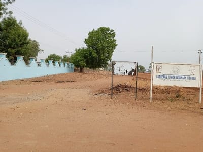 The image shows a dirt road in front of a blue wall with white trim. There is a sign on the right side of the road that reads "LUTHERAN JUNIOR SEMINARY, IMBAMBA." There is a small tree to the left of the sign and another large tree further back on the left side of the image. The road leads to a clearing with a few trees in the distance. The image was taken in the city of Rumde Waziri, Nigeria, at the coordinates latitude: 9.188, longitude: 12.525.
