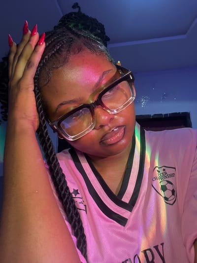 An individual, identified as a female, is depicted in a medium close-up portrait within an indoor setting in Ipetumodu, Nigeria. The subject possesses dark skin, is wearing large, clear-framed glasses with dark temples, and has braided dark hair styled upwards. Her right hand, adorned with long, pointed red acrylic nails, rests on her head near the hairline. She is wearing a pink V-neck jersey featuring black stripes on the collar and shoulders, and a "SOCCER CHAMPIONSHIP" logo with a soccer ball on the upper right chest area. A distinct rainbow light effect, along with pink, blue, and green ambient lighting, illuminates her face and upper torso, casting a prominent pink highlight on her forehead and glistening her lips. The background is indistinct, appearing as a dark blue surface.