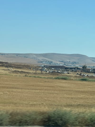 The image depicts a landscape in the city of al-Qasoum Regional Council, Israel. A light blue sky takes up the left portion of the frame. Adjacent to the sky is a dry, mountainous terrain with a town nestled along its base. The town consists of low-lying buildings. To the right of the town and mountain base stretches a field of dry, yellow grass. Some green vegetation is also visible, mainly on the far right edge of the frame. There is a fence line between the town and the field.