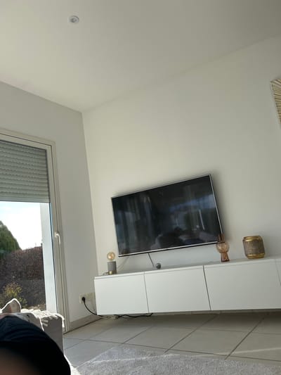The image depicts an indoor residential space, likely a living room, located in Veauchette, France. The room features white walls and a white ceiling with a single circular recessed light fixture. A large, dark-screened flat-panel television is mounted centrally on a wall. Below the television, a white floating media console, comprising three cabinet units, is affixed to the wall.
Upon the console, from left to right, are: a decorative lamp with a grey concrete-like base, a brass-colored accent, and an exposed filament bulb; a small dark spherical object; an amber-colored glass vase with a textured surface; and a cylindrical, gold-colored woven decorative object. Reflections on the television screen show elements of the room, including what appears to be a light-colored sofa. A partial view of a decorative wall hanging with a sunburst pattern is visible on the upper right wall.
On the left side of the room, a large white-framed glass door or window is partially open or closed. A grey, horizontally slatted roller shutter is partially retracted at the top of this opening. Sunlight streams into the room through this window, illuminating the tiled floor and a light-colored, shaggy rug in the foreground. Outside the window, green foliage and brown leafless bushes are visible under a bright sky. A white electrical outlet with a black cord plugged in is situated on the wall below the window frame. In the lower-left foreground, a human leg, clothed in dark fabric and light-colored socks, rests on a light-colored textile, presumably part of a sofa or chair. No other individuals or clear actions are discernible.