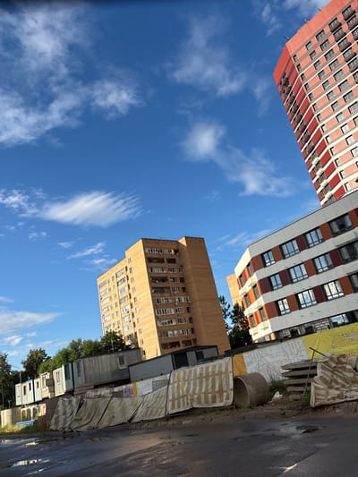 The image captures an upward-looking view of several tall buildings against a bright blue sky with scattered white clouds, suggesting a clear, sunny day, likely in the afternoon. The buildings are diverse in their architecture and materials.

On the left, a predominantly red and white building with many windows is seen. To its right, a modern building with a white facade is interspersed with horizontal bands of reddish-brown and orange, also featuring numerous windows. In the lower half of the image, a lighter brown, brick-like building with many windows is visible, appearing older than the other two. Next to this, there are several smaller, corrugated metal structures that resemble portable cabins or construction site offices, lined up along a street. Some of these are partially covered by tarps with a checkered pattern.

The foreground on the right shows a wet asphalt road, indicating recent rain or that the area is prone to puddles. There are also some construction materials visible, including a large concrete pipe and rolled-up sheeting, suggesting that some form of construction or renovation is taking place. Lush green trees are visible behind the smaller structures, contrasting with the urban environment.

No people are clearly visible in the frame. The overall scene depicts an urban environment with residential buildings and indications of ongoing construction or development. The perspective emphasizes the scale of the buildings against the sky. No legible text is discernible in the image.