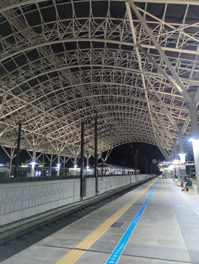 The image shows a train platform at Singyeongju Station in Gyeongju-si, South Korea, at night. The platform is illuminated by overhead lights.  The distinctive architecture is a large, curved, white steel framework covering the platform. A tactile paving strip and a blue line with Korean writing run along the platform edge. People are sitting and standing on the platform in the distance.