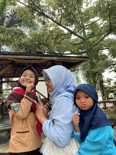 Three individuals are depicted outdoors in Kalitengah, Indonesia. In the center, an adult female with a light blue patterned hijab, light blue long-sleeved shirt, and white skirt holds a smartphone, looking towards it. To her left, an older female child in a brown scout-style uniform with a red and white neckerchief and a sleeve badge smiles, holding a thin stick to her mouth, and wears a dark brown hijab. To the adult's right, a younger female child in a dark blue hijab and light blue long-sleeved shirt holds a skewer with a red, flower-shaped food item near her mouth, looking directly forward. Behind them, a bamboo gazebo structure is visible. The background features trees with light green and white variegated leaves, with a palm tree on the far left. Wooden outdoor furniture is partially visible in the distant right background.