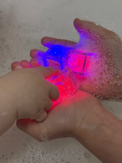 The scene is captured indoors, specifically in a bathroom in Rusalevka, Russia, depicting two hands interacting with luminous objects in a body of water covered with white foam. An adult's right hand, palm upward, supports three square, transparent light-emitting cubes. One cube on the left emits a vivid blue light, displaying faint white text indicating "ICE CUBE" and "MADE IN CHINA". Adjacent to it, a cube emits a bright pink light, and another below it emits a red light. The adult hand is partially submerged, covered with water droplets and foam, particularly on the knuckles and wrist. A child's left hand, with plump fingers, reaches from the left side of the frame to gently touch the red-glowing cube. The child's hand is also wet and appears to have a few water droplets. The water surface, which forms the background, is entirely covered in small to medium-sized white foam bubbles, suggesting a bath or wash basin setting. The lighting emanates primarily from the glowing cubes, casting colored light onto the skin of both hands and the surrounding foam.