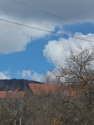 The image shows a view of Gintsi, Bulgaria. The sky is partially cloudy, with a mix of blue sky and gray clouds. A building with a red tile roof is partially visible on the right side of the frame, obscured by bare trees. Utility lines are seen on the left against the sky.