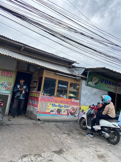 The image shows a street scene in Katapang, Indonesia (latitude: -6.997, longitude: 107.554). A man wearing a black hooded sweatshirt and blue jeans is standing in front of a small storefront. The storefront has a red and yellow sign that says "MONGGO KAMPIR BASO RUDAL & BASO BERANAK "Ilas Baje Solo"". The sign lists different types of bakso (meatballs), including bakso rudal, bakso urat, bakso mercon, bakso telor, bakso cincang, mie ayam, and mie letok. The man is looking at his phone. A motorcycle is parked in the street to the right of the storefront. The motorcycle has a blue helmet on the seat. A person wearing a tan jacket is sitting on the motorcycle. The motorcycle has a "Honda" logo on the side. There are many power lines running across the sky. The storefront is next to another building that has a green sign with the words "PONDOK DURIAN" on it. The sign also says "DURIAN MONJONG SEDAP" and "KETAN". The buildings appear to be made of concrete. The street is paved. The sky is overcast. The image is taken from street level. The scene is set in an urban environment with an active street and businesses.