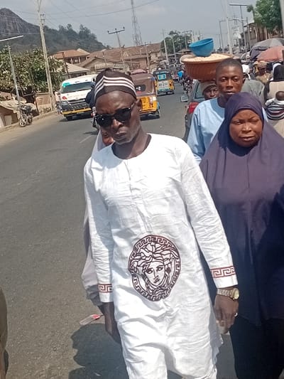 Three people are the focus on a sunny city street. One man wears a white, long-sleeved shirt with a decorative Medusa-like emblem and an ornate pattern on the cuffs, accessorized with sunglasses, a striped cap, and a gold wristwatch. Behind him, a man in a light blue shirt and a woman in a dark blue head covering are visible. The background includes buildings, power lines, a white truck, yellow tricycle taxis, and a mountain in the distance.
