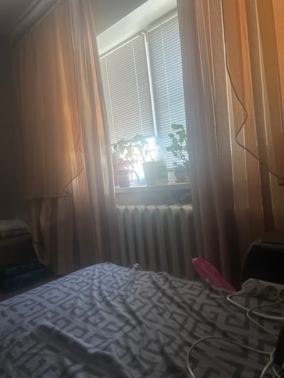 The image depicts an indoor scene, likely a bedroom, with a window in the background that is covered by horizontal blinds. On the windowsill, several potted plants are visible. The room is sparsely furnished, with a radiator located beneath the window. A bed with patterned grey bedding occupies the foreground of the image. To the left of the bed, a stack of folded cloths rests on a surface. To the right of the bed, an electrical extension cord with multiple sockets and plugs is situated. The overall lighting suggests it is daytime, with sunlight filtering through the window and curtains. The image does not contain information that would allow for definitive placement in the city of Sinegorye, Russia.