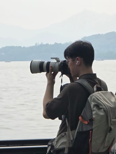 A man stands on a walkway, holding a large camera with a telephoto lens to his eye, looking out over a body of water towards hazy mountains. He is wearing a dark shirt and a backpack with grey and orange accents. The water appears calm, with a few small boats visible in the distance. The overall atmosphere is muted, suggesting an overcast or foggy day. The scene takes place in Shangcheng District, China.