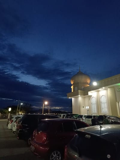 In the city of Mee Tanjong Usi, Indonesia, a mosque with a golden dome stands illuminated against a deep blue twilight sky filled with dark clouds. The mosque features decorative carvings and arched windows. In front of the mosque, a parking lot is filled with several cars, including a red hatchback and black SUVs. A few individuals are visible near the vehicles. Streetlights are also present, casting a warm glow in the dim light.