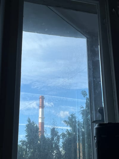 The image depicts a view through a window of a smokestack with alternating red and white bands, likely a power plant or industrial facility, situated in the city of Vsevolozhsk, Russia. The smokestack is tall and stands against a blue sky with scattered white clouds and distinct contrails from aircraft. In the foreground, partially obscuring the view, are the green leaves of trees. The window frame is visible on the sides of the image, with the right side showing a dark object, possibly part of a device or furniture, and a window latch. The glass of the window appears somewhat smudged.