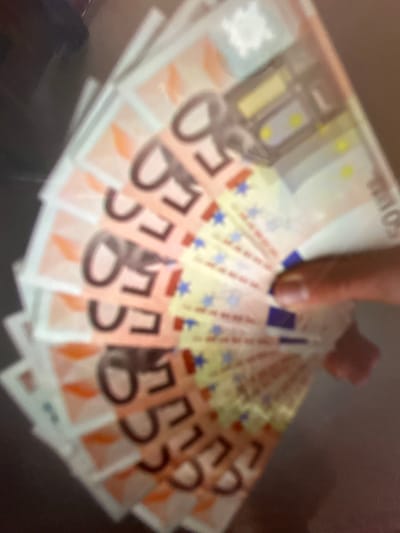The image shows a close-up, blurred view of a fan of fifty euro banknotes. A thumb is holding the banknotes from the right side of the image. The background is dark and out of focus, with some indistinct shapes. There is no discernible information about the location in Gelendzhik, Russia, as the image is purely a close-up of currency.