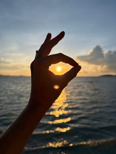 A silhouette of a hand forms a circle around the sun during sunset over the ocean at Baan Plai Laem, Thailand. The sun is a bright orb, partially obscured by the thumb and index finger. The ocean surface reflects the golden light of the setting sun, creating a shimmering effect. The sky is a gradient of blue and orange, with scattered clouds. The image captures a moment of serene natural beauty.