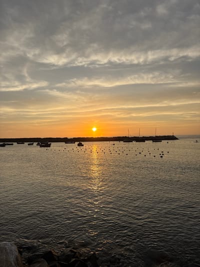 The image captures a sunset scene in Chorrillos, Peru. The sun is positioned near the horizon, casting a golden glow across the water and sky. The sky features layers of clouds, with the lower clouds displaying a greyish-white hue and the upper clouds tinged with orange and yellow. A line of moored boats extends along the coastline. The water reflects the sunlight. The edge of a rocky shoreline is visible in the bottom right corner of the image.