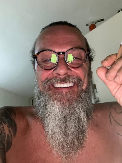 A shirtless man with a full grey beard and a ponytail is looking directly at the camera and smiling. He is wearing round, tortoise-shell glasses. His right hand is raised, holding what appears to be a cigarette or a joint between his thumb and index finger. The background is a plain, lightly colored wall, with a portion of what appears to be a shelf visible at the top right, with an orange object that looks like a butane torch resting on it. The lighting suggests an indoor setting. The image is a close-up selfie. The person's location is unknown.