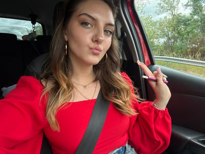 A person is shown from an unusual upside-down perspective in what appears to be the interior of a car. The individual is wearing a red, long-sleeved top and a seatbelt. They have light brown hair with blonde highlights, and their nails are painted with a multi-colored design. They are holding a small purple device in their hand. The background features a glimpse of trees through the car window and parts of the car's interior. There is nothing to indicate that the image was taken in the city of Bliznets, Bulgaria.