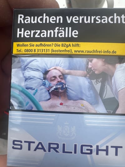 This is a close-up, vertical shot of a pack of cigarettes in Krefeld, Germany. The packaging displays a stark warning message in German: "Rauchen verursacht Herzanfälle" which translates to "Smoking causes heart attacks." Below this, it offers help for quitting with "Wollen Sie aufhören? Die BZgA hilft:" meaning "Do you want to quit? The BZgA helps:" followed by a phone number and website address.
The dominant visual on the pack is a photograph of a person in a hospital bed, appearing unwell and connected to medical equipment, including a ventilator. Another person, likely a visitor, is shown leaning over the patient with a somber expression. The overall mood conveyed is serious and cautionary, emphasizing the severe health consequences of smoking. The right side of the pack features the brand name "STARLIGHT" in large, bold letters. The lighting appears to be artificial, consistent with an indoor, likely medical, setting. The image is well-lit, allowing for clear readability of the text and details within the photograph.