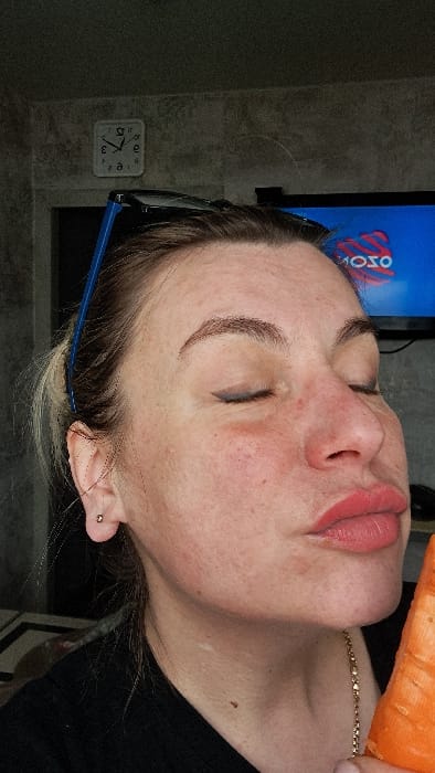 The scene is an indoor domestic setting in Bel'kovo, Russia. A female subject, of apparent adult age, is captured in a mid-shot, her head tilted slightly, with her face in profile, directed towards the lower right. Her eyes are closed, with visible black winged eyeliner. Her lips are pursed and colored with a light red lipstick, positioned in close proximity to a large, orange, raw carrot held upright in her right hand, which is mostly out of frame. She exhibits fair skin with visible pores, minor redness on the cheeks, and well-defined eyebrows. Blue-framed glasses are resting on top of her head, and a small gold-colored stud earring is visible in her left earlobe. A thin gold-colored chain is worn around her neck, partially visible over a dark, indeterminate garment. The background consists of a dark, textured wall. To the upper left, a white square wall clock with black hands displays a time approximating 11:45 or 12:45. To the upper right, a television screen is visible, displaying a blue background with a white and red graphic, possibly a logo, which appears as "MOTO" or "OZO" in reverse.