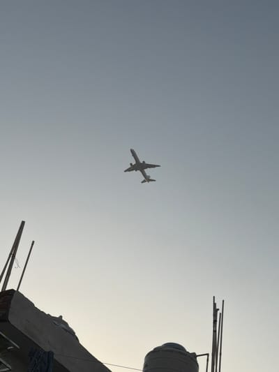 A distant silhouette of a passenger airplane is seen flying in the hazy sky, positioned slightly above the center of the frame. In the foreground, the upper edge of a rooftop structure is visible on the left, characterized by exposed bricks and a sloping concrete surface. On the right side of the foreground, a water tank and several thin vertical poles, possibly antennas or structural elements, are present. The scene appears to be from an urban environment, likely a rooftop in Delhi, India, given the context. The lighting suggests it is either early morning or late afternoon, with a bright, overcast sky.