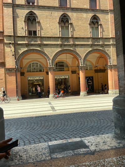 The image shows a street scene in Bologna, Italy, featuring an overhead view of a colonnaded walkway and the facade of a building housing a "Pull & Bear" store. The building has arched entryways and windows. People are present, some walking along the walkway and others appearing to be near the store entrance. Bicycles are visible in the street. The street is paved with cobblestones. The photo is taken from an elevated perspective, possibly from within another building or structure overlooking the street. The natural light casts shadows on the building and the street.