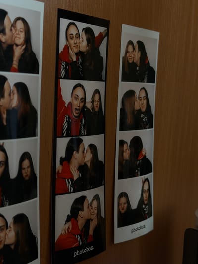 The image shows three strips of photobooth pictures laying on a wooden surface. The photobooth strips depict two individuals, presumably a couple, interacting in various poses. Some frames show them kissing or embracing. In one frame, one individual makes bunny ears behind the other's head. The strips are labeled "photobox" along the right edge. The location of the image, Shul'gino, Russia, is not depicted in the picture.