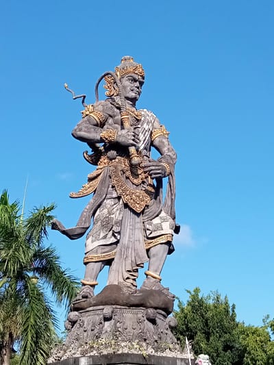 A low-angle, full-shot photograph shows a large stone statue of a Balinese deity or warrior, predominantly gray with gold accents, standing against a clear, vibrant blue sky. The statue, elaborately adorned with intricate carvings and golden decorative elements, holds a weapon in its hands. The figure wears a headdress and ornate clothing, with a rope coiled over its shoulder. The statue stands on a detailed stone pedestal adorned with decorative carvings. To the left of the statue, a palm tree with lush green fronds is partially visible, and to the right, a dense green canopy of trees forms the background. The overall impression is one of grandeur and cultural significance, likely located in Blahbatuh, Indonesia, given the prominent Balinese artistic style.