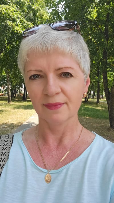 An outdoor selfie of a Caucasian woman with short, gray hair and sunglasses pushed onto her head. She is wearing a light blue shirt and a gold necklace with a pendant. Her makeup includes pink lipstick and eyeliner. The background shows a park with trees and a paved walkway, suggesting a sunny day in Belaazyorsk, Belarus.