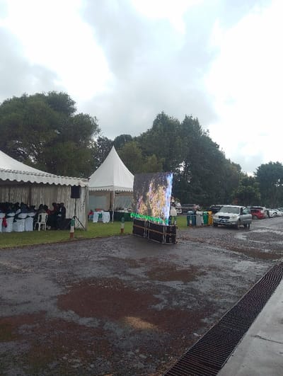 This image captures an outdoor event taking place in Gitaru, Kenya, under an overcast, cloudy sky. The ground is wet and appears to be a mix of gravel and mud with puddles, suggesting recent rainfall.
On the left side of the frame, a large white rectangular marquee is erected, under which numerous people are seated on rows of white chairs, some decorated with red sashes. They appear to be attending a gathering or presentation. A large black speaker on a tripod stand is positioned in front of this tent. Adjacent to the main marquee, a smaller, pointed white canopy tent stands alone.
Centrally located is a prominent, large, vertical LED screen displaying what looks like a vibrant natural scene, possibly trees or foliage. The screen is mounted on a dark, multi-layered base, which might house associated equipment.
To the right of the LED screen, several cars are parked on the wet, gravelly surface, including a silver SUV. Near the parked cars, a cluster of colorful waste bins (green, blue, yellow) is visible. A long, dark metal drainage grate extends along the bottom right edge of the frame. Small red and white striped poles are intermittently placed on the ground, possibly marking boundaries or pathways.
The scene depicts an organized outdoor event, equipped with temporary structures for shelter and seating, as well as a large screen for visual presentations. The damp conditions are a notable aspect of the setting. No discernible text is visible in the image.