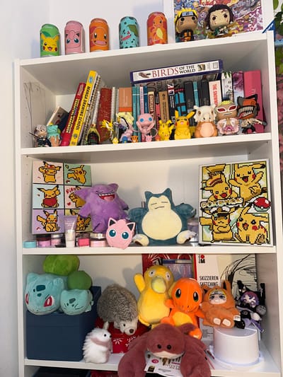 A white shelving unit is filled with an assortment of items, primarily Pokémon-themed merchandise and books.
The left shelf contains books stacked vertically, including "Birds of the World" and "Manga Drawing School," along with manga books and trinkets such as figurines. Pokemon themed cans are placed vertically on the side of this shelf.
The middle shelf features a framed picture composed of individual square units depicting various Pikachu poses. A plush Snorlax and a Gengar sit below the framed artwork, with additional smaller items and makeup positioned near the bottom.
The right shelf is filled with stuffed toys, including Pokemon like Charmander and Psyduck, interspersed with other items such as a hedgehog plush, a small white bunny, and a pair of Bulbasaur plushies. A blue storage box is also in the rightmost shelf.
The environment shows a clean, well-lit space. There are no people visible in the image. There is no indication that this is in Ljubljana, Slovenia.
