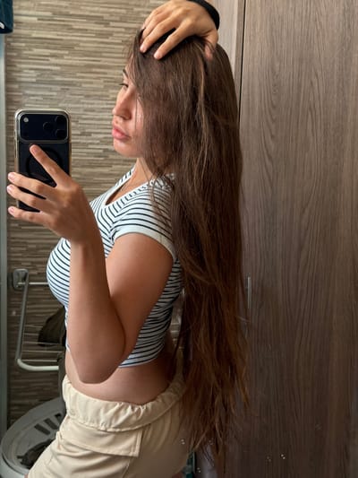 A young woman with long, brown hair is standing in front of a wooden closet and a tiled wall, taking a selfie with her phone. She is wearing a striped t-shirt and light-colored shorts. Her hair is pulled back slightly with her hand. The location appears to be a domestic interior.