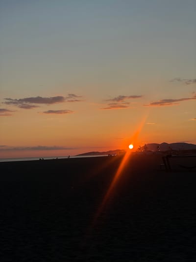 A wide shot captures the sunset on a beach in Donji Štoj, Montenegro. The sky transitions from a pale blue at the top to warm oranges and pinks near the horizon. A few scattered clouds are tinged with color from the setting sun. The sun itself is a bright, glowing orb, casting a strong beam of light across the scene. Silhouetted figures of people are visible on the dark sand of the beach, some sitting and others standing. In the distance, faint outlines of hills and some structures suggest a coastal town or resort. The foreground is dominated by the dark sand of the beach, with some texture indicating ripples or dunes. A hammock and a bench are visible in silhouette on the right side of the frame. The overall impression is a serene evening at the beach.